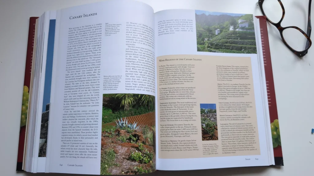 Un libro de vino abierto por la página de las Islas Canarias, mostrando fotos de sus viñedos únicos y texto sobre la región. Unas gafas de lectura están cerca.