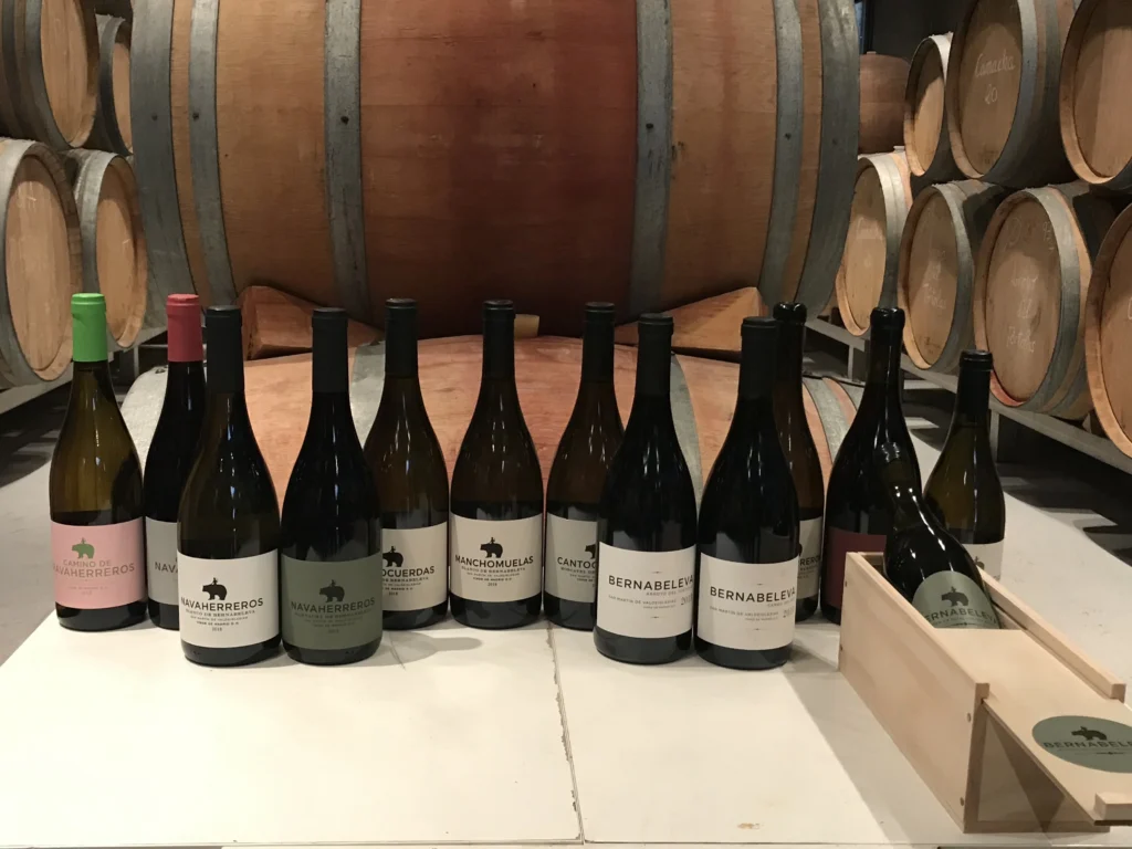 Colección de botellas de vino de la bodega Bernabeleva, incluyendo Navaherreros y Cantocuerdas, dispuestas frente a barricas de roble en una bodega.