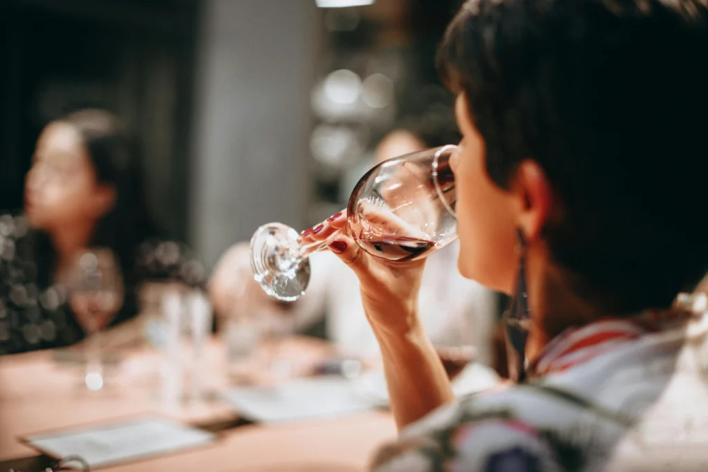 Mujer catando una copa de vino tinto, observando su color y oliendo sus aromas en un evento de degustación con más gente al fondo.