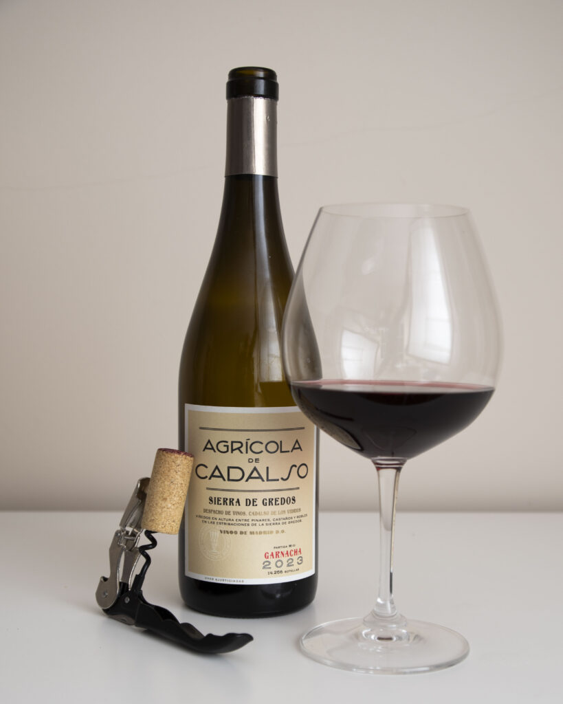 Botella de vino tinto Agrícola de Cadalso Sierra de Gredos Garnacha 2023 junto a una copa de vino llena y un sacacorchos con el corcho.