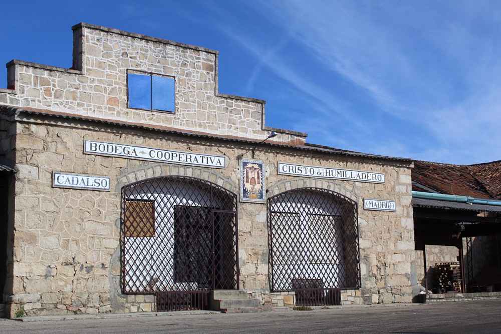 Vista frontal de la Bodega Cooperativa Cristo del Humilladero de Cadalso de los Vidrios, con su fachada de piedra, rejas en las puertas y carteles de 'Bodega Cooperativa', 'Cristo del Humilladero', 'Cadalso' y 'Madrid'.