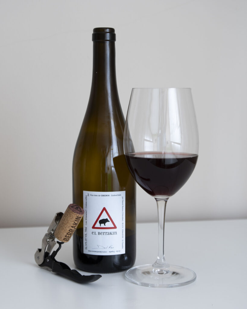 Botella de vino tinto 'El Berrakín 2023' de Daniel Ramos, con etiqueta blanca y un dibujo de un verraco, junto a una copa de vino tinto servida y un sacacorchos.