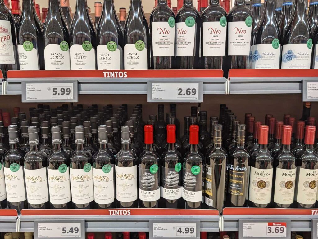 Estantería de supermercado con múltiples botellas de vino de distintas marcas (Finca La Cruz, Neo, TRAMUZ) de Ribera del Duero y Valdepeñas con sellos de 90 y 91 puntos.