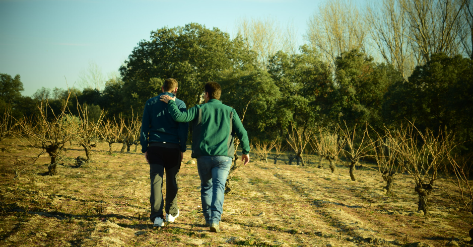 Dos hombres de espaldas, vestidos con chaquetas, caminando por un viñedo podado en vaso en invierno, con árboles boscosos al fondo.