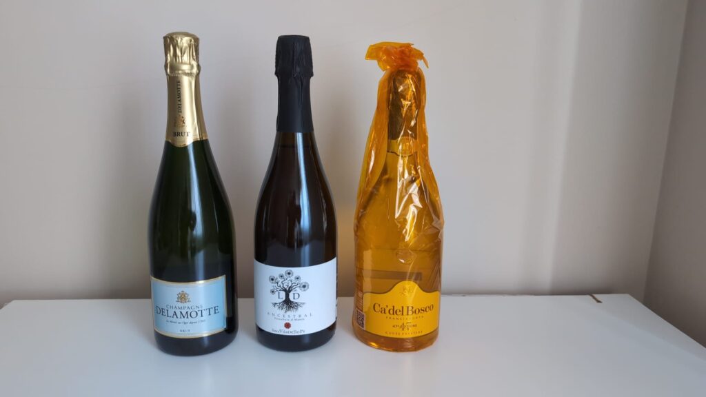 Tres botellas de espumosos de diferentes regiones: Champagne Delamotte, una botella de Cava Ancestral y una botella envuelta en papel dorado de Franciacorta Ca' del Bosco.