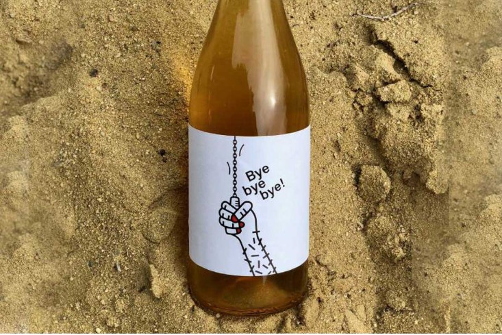 Botella de vino ámbar con etiqueta ilustrada “Bye bye bye!” colocada sobre arena.