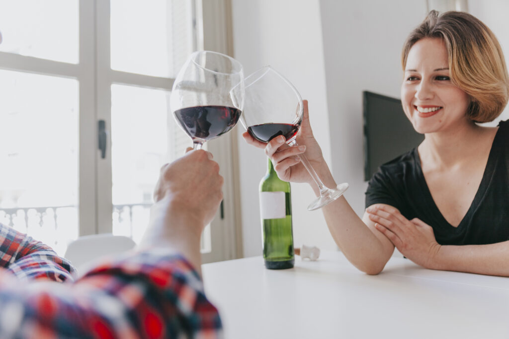 Dos personas brindando con copas de vino tinto sentadas a una mesa junto a una ventana.