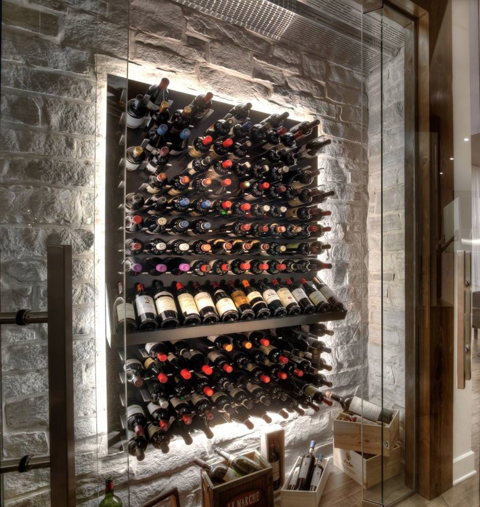 Bodega moderna con botellas de vino almacenadas en horizontal en una pared iluminada tras un cerramiento de cristal.
